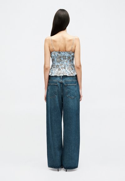 Femme aux longs cheveux noirs portant un haut sans manches bleu à fleurs et un jean bleu à jambes larges, debout face à un fond blanc uni.