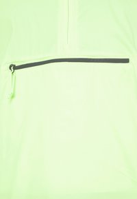 Chaqueta cortavientos verde claro con medio cierre frontal y un bolsillo negro con cremallera. Fabricada en un material suave y ligero.