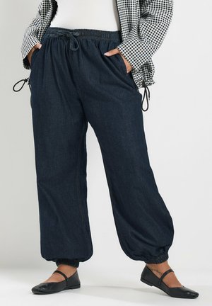 Persoon draagt donkere losse broek met elastische boord, gecombineerd met zwarte platte schoenen en een zwart-wit geruite top met lange mouwen.
