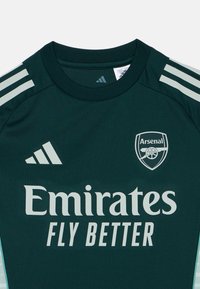 Ciemnozielona koszulka piłkarska z krótkim rękawem, z jasnoszarymi paskami, logo Arsenalu i napisem "Emirates Fly Better".