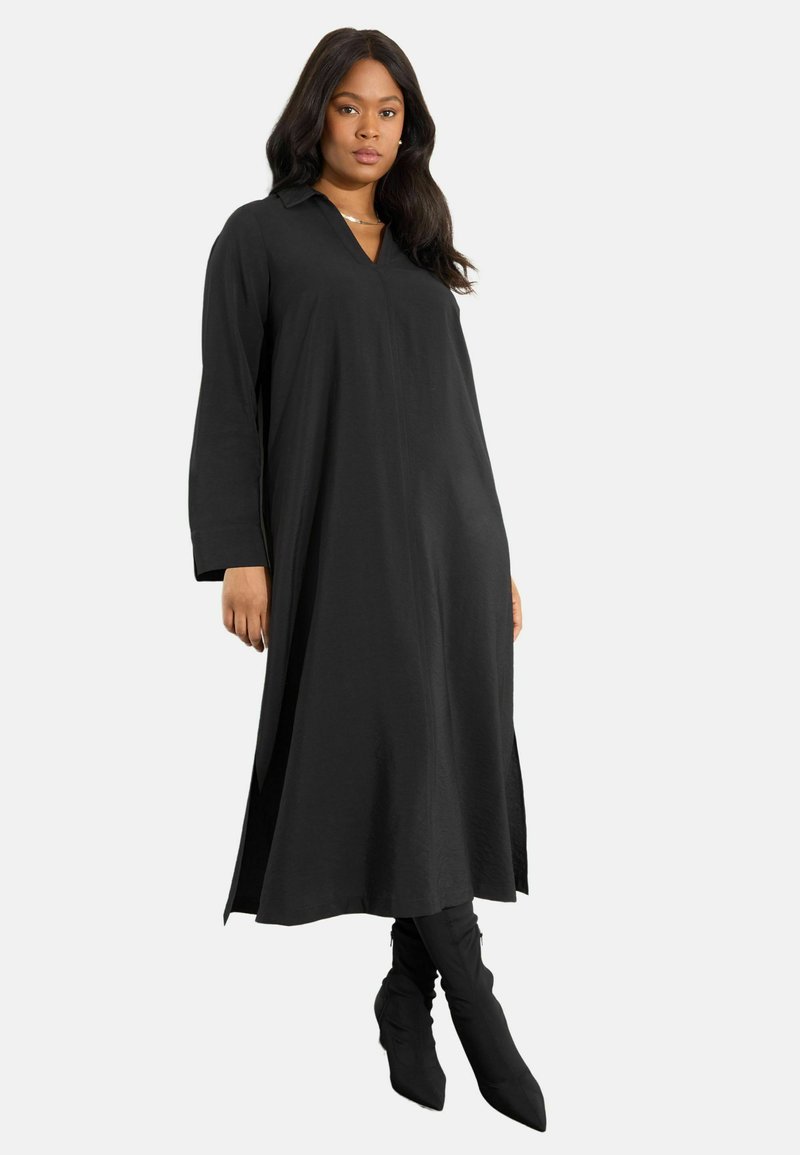 Schwarzes langärmliges Kleid mit einem kragenlosen V-Ausschnitt, lockerer Passform und seitlichen Schlitzen, kombiniert mit schwarzen Stiefeln bis zum Knie.