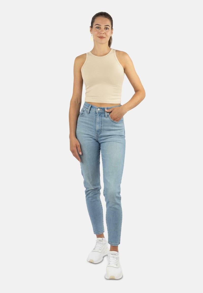 Eine Frau steht mit einem beigen, ärmellosen Crop-Top, hellblauen, hochtaillierten Jeans, weißen Sneakers und goldenen Ohrringen da, mit einer Hand in der Tasche.