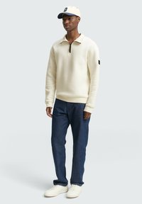 Crème zip-hals trui met een gestructureerd patroon, gecombineerd met donkerdenim jeans en witte sneakers. De outfit wordt aangevuld met een beige en marineblauwe pet.