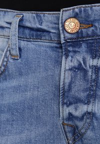 Jasnoniebieskie dżinsy z denimu z miedzianym zapięciem na guzik, akcentami w postaci szwów oraz charakterystycznym wzorem kieszeni z wyblakłymi obszarami i fakturą.