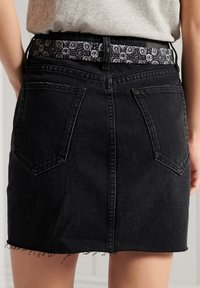 Falda de mezclilla negra con un dobladillo deshilachado, que presenta dos bolsillos traseros y un cinturón de patrón en blanco y negro visible en la cintura.