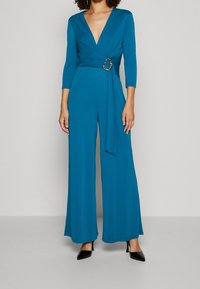 Combinaison teal avec un décolleté en V profond, des manches longues, un devant croisé et une taille cintrée avec un détail décoratif en anneau doré. Jambes évasées.