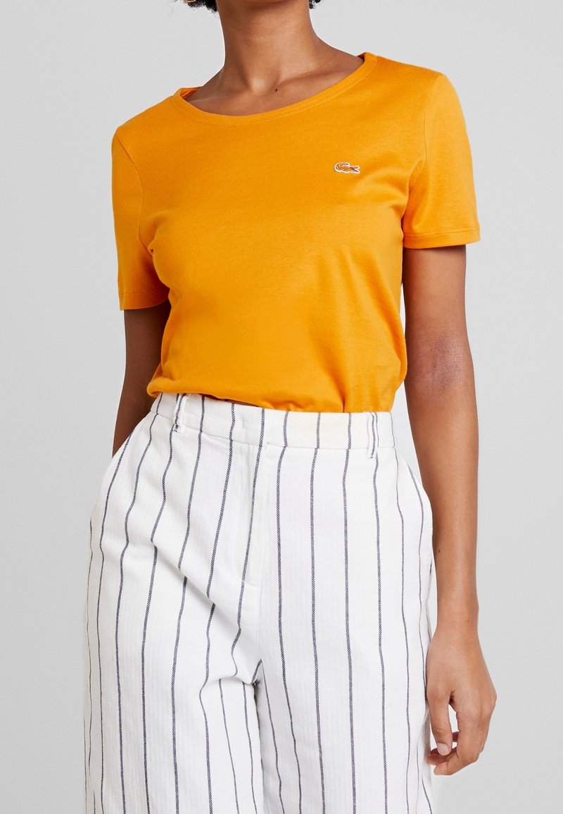 T-shirt en coton orange avec un col rond et des manches courtes, ornée d'un petit logo brodé sur la poitrine, associée à un pantalon blanc à rayures.