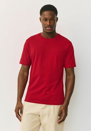 Man met kort krullend haar die een effen rood T-shirt met korte mouwen en beige broek draagt, staand tegen een effen lichte achtergrond.