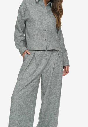 Frau trägt ein graues, langärmeliges Hemd mit Knöpfen und weite, plissierte graue Hose, mit einer Hand in der Tasche, stehend vor einem weißen Hintergrund.