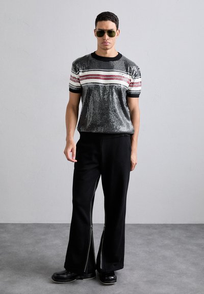 Pull à manches courtes en sequins noirs et argent, avec des rayures rouges et blanches, associé à un pantalon ample noir avec des détails zippés.