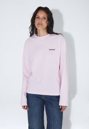 Jonge vrouw met krullend donker haar, draagt een lichtroze shirt met lange mouwen met het "Dr.Denim" logo en een blauwe spijkerbroek, staand tegen een witte muur.