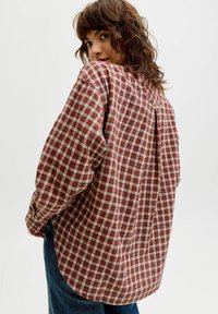 Karirana srajca v rdečih, bež in modrih odtenkih. Ohlapen kroj z dolgimi rokavi, ukrivljenim robom in naborkom na zadnji strani. Iz bombažnega materiala.