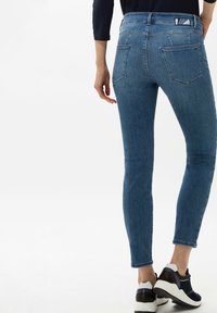 Dünne blaue Jeans aus Denim mit glatter Textur, ausgestattet mit zwei Gesäßtaschen und genähten Details, kombiniert mit schwarz-weißen Sneakers.