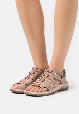 Füße in beigen Outdoor-Sandalen mit roten Schnürsenkelschlaufen und robusten Gummisohlen auf einem weißen Hintergrund.
