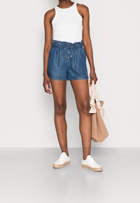 Denim-Shorts mit elastischem Bund und Kordelzug, mit lockerem Sitz, Seitentaschen und einem Fransenabschluss. Kombiniert mit einem weißen Tanktop.