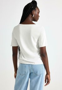 T-shirt bianco a maniche corte con scollatura quadrata, realizzato in tessuto morbido. Abbinato a jeans a vita alta di colore azzurro chiaro. Dettaglio di treccia evidente nei capelli.
