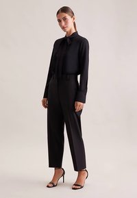 Schwarze Bluse mit Schleifenkragen und langen Ärmeln, kombiniert mit hoch taillierter schwarzer Hose. Glatter Stoff und taillierte Passform, ergänzt durch schwarze Absätze.