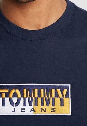Mężczyzna w granatowej koszulce z okrągłym dekoltem i prostokątną naszywką z logo "Tommy Jeans" w kolorach białym, żółtym i granatowym na klatce piersiowej.