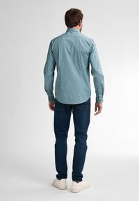 Camicia turchese a fantasia con colletto button-down, maniche lunghe e orlo arrotondato; abbinata a jeans blu scuro e sneakers bianche.