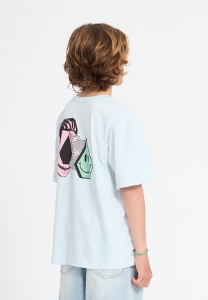 Enfant aux cheveux bruns mi-longs portant un T-shirt bleu clair surdimensionné avec un imprimé géométrique pastel et un visage souriant au dos.