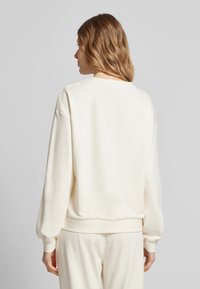 Felpa color crema con vestibilità rilassata, spalle scese e polsini a costina. Tessuto morbido con una texture liscia, design minimalista.