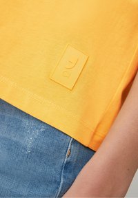 Zbliżenie na żółty brzeg koszulki z haftowaną naszywką logo, w połączeniu z niebieskimi dżinsami i nagą ręką.