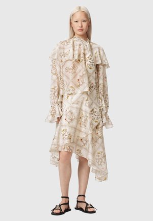 Femme portant une robe asymétrique crème à motifs avec des volants et des manches longues, associée à des sandales noires plates avec des brides à la cheville.