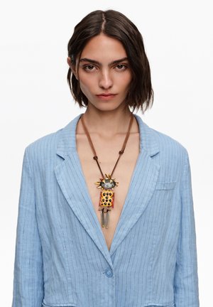 Kobieta o krótkich ciemnych włosach nosi otwartą, jasnoniebieską marynarkę w prążki oraz kolorowy, nietypowy naszyjnik z mieszanymi materiałami.