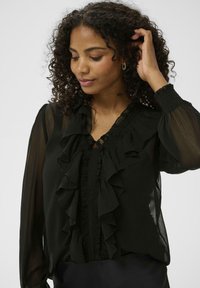 Zwarte, doorschijnende blouse met lange, gebloemde mouwen, ruches aan de voorkant en een V-halslijn. De stof lijkt lichtgewicht met aangehechte ruches.