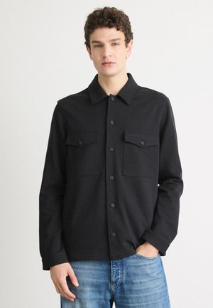 Black button-up jacket with a collared design, two front pockets, and a textured fabric. Worn over blue denim jeans.