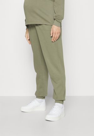 Lower body of a person wearing olive green jogger pants, matching sweatshirt, white socks, and white sneakers, standing on a light gray floor.