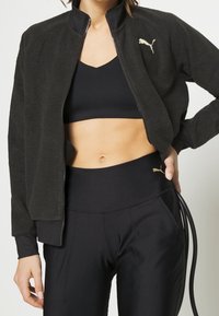 Czarna polarowa kurtka z zamkiem z przodu i wysokim kołnierzem, zdobiona złotym logo. Noszona na czarnym sportowym biustonoszu i legginsach z wysokim stanem.