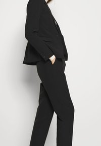 Completo nero su misura con un blazer elegante con rever a scollo e pantaloni a gamba dritta. Il tessuto ha una trama liscia e un design minimalista.