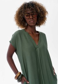Zielona, lekka sukienka z krótkim rękawem, dekoltem w kształcie litery V i luźnym krojem. Materiał ma gładką fakturę, podkreśloną dużymi bransoletkami na nadgarstku.