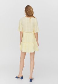 Gelbes kurzes ärmelloses Kleid mit einem gestuften Design und Spitzenakzenten entlang der Taille, das eine lockere Passform und einen leichten Stoff aufweist.