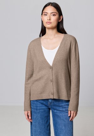 Cardigan beige avec un col en V, fermeture à boutons et ourlet côtelé. Porté par-dessus un top blanc, associé à un jean bleu. Texture lisse et coupe décontractée.