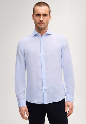 Man staat recht vooruit kijkend, draagt een lichtblauw overhemd met lange mouwen en een donkere broek tegen een effen achtergrond.