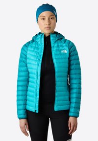 Teal jacka med vadderad textur, huva, och logotyp på vänster bröst. Bärs över en svart topp, tillsammans med svarta byxor.