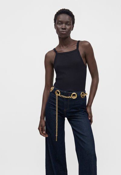 Kvinde iført sort ærmeløs top, mørke vidde jeans med høj talje og stor guldkæde som bælte, stående mod ensfarvet lys baggrund.