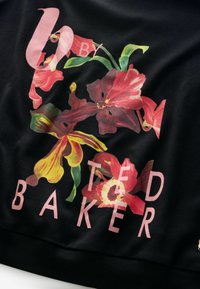 Fekete pulóver élénk piros, sárga és zöld virágmintával, rajta világos rózsaszín betűkkel a "U BY TED BAKER" felirat.