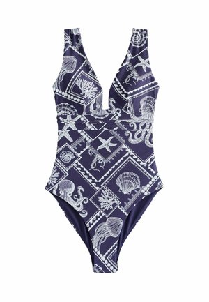 Nő kék fürdőruhában, fehér tengeri lényekkel és kagylómintákkal, egy víz test közelében áll, tiszta ég alatt.