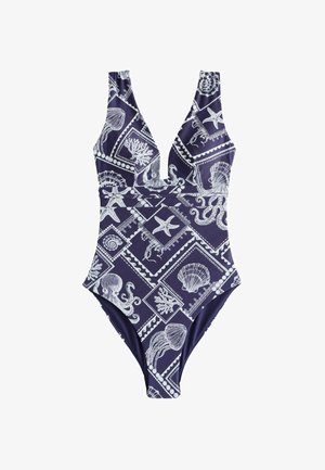 Vrouw draagt een blauw badpak met witte zeedieren en schelpenpatronen, staand bij een wateroppervlak onder een heldere lucht.