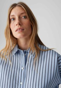 Chemise rayée bleu clair avec col classique, boutonnée sur le devant et manches courtes. Le tissu présente une texture lisse avec un léger éclat.