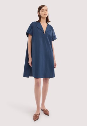 Kobieta stojąca w luźnej, sięgającej kolan granatowej sukience z krótkimi rękawami, dekoltem w szpic oraz brązowych wsuwanych butach na szpic ze złotym detalem.