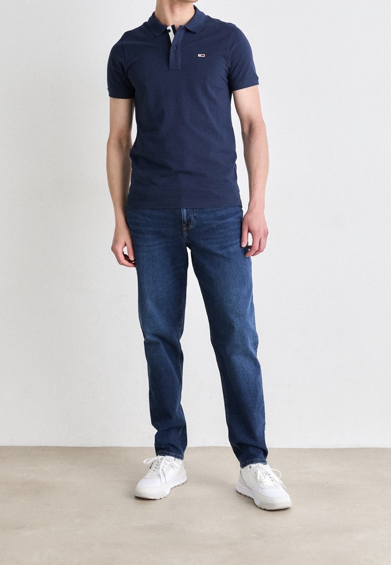 Polo-shirt bleu marine avec un col à trois boutons, accents blancs et rouges, associé à un jean bleu et des baskets blanches. Pose debout directe.