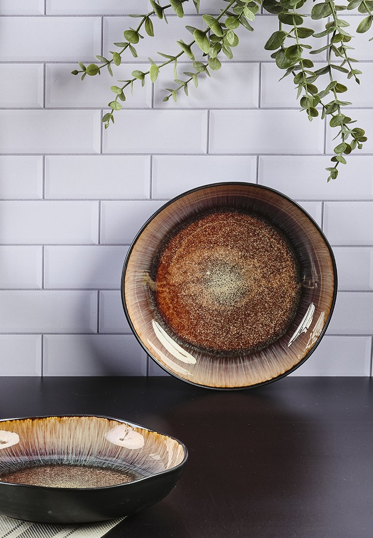 Zwei Keramikschalen mit glänzender Oberfläche; eine größere und runde, die einen Farbverlauf von Brauntönen zu Goldtönen aufweist, und eine kleinere, oval geformte.