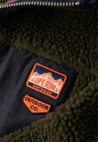 Grünes Fleece-Innenfutter mit Zickzack-Stich, mit orange bestickten Aufnähern auf schwarzem Stoff, die Berg- und Markenlogos zeigen.