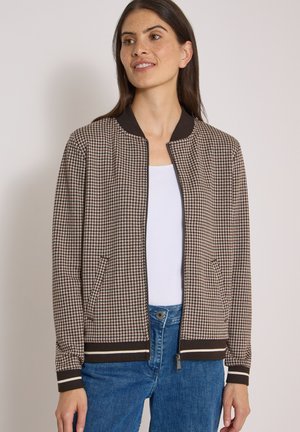 Femme portant une veste zippée marron et blanche à motif pied-de-poule, par-dessus un haut blanc et un jean bleu, regardant sur le côté avec un léger sourire.