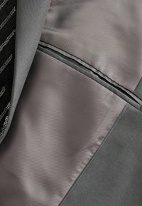Gray suit jacket interior featuring a smooth gray lining, contrasting textured black fabric, and a horizontal pocket along the seam.