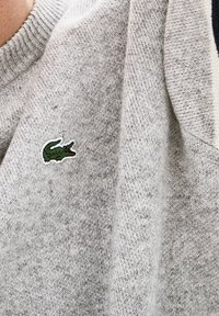 Šedý pletený svetr s žebrovaným výstřihem a malým zeleno-bílým krokodýlím logem na hrudi. Jemná textura s melírovaným vzorem.